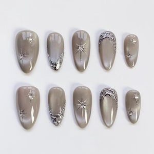 May include: Set of ten almond-shaped, press-on nails in a neutral taupe color. Some nails feature silver starburst designs or ornate silver accents. The nails are arranged on a white surface, showcasing a modern nail art design.