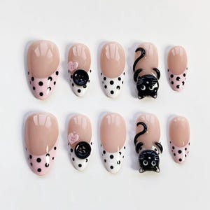 Puede incluir: Un conjunto de diez uñas postizas ovaladas. Los diseños incluyen una base rosa con lunares negros, una base blanca con lunares negros, un corazón rosa y un botón negro, y un diseño de gato negro.