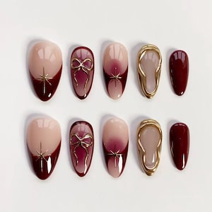 May include: A set of ten artificial nails in various designs. The nails feature a nude base with burgundy tips, gold starburst accents, gold bow details, and gold-framed designs. One nail is solid burgundy.