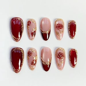 Pode incluir: Conjunto de dez unhas artificiais em várias formas. As unhas apresentam um esquema de cores bordô e dourado. Algumas unhas têm um contorno dourado, enquanto outras têm um desenho de rosa dourada. As unhas estão dispostas sobre uma superfície branca.