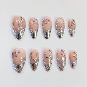 May include: Set of ten artificial nails in various sizes. Each nail features a nude-colored base with a silver metallic design. The silver design includes a swirl pattern and a metallic tip, creating a decorative and elegant look.