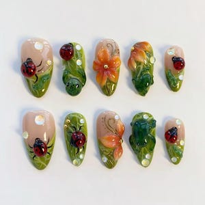 May include: A set of ten artificial nails with a nature theme. The nails feature ladybugs, flowers, and leaves in various designs. Colours include red, green, orange, and beige. Some nails have a glossy finish with small, clear embellishments.