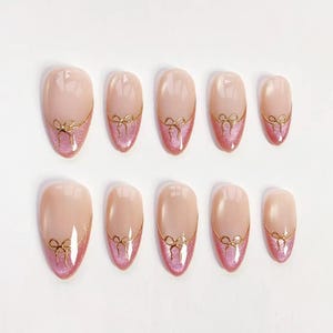 May include: A set of ten artificial nails in various sizes. Each nail features a nude base with a pink glitter tip and a gold bow design. The nails are oval-shaped and arranged on a white surface.
