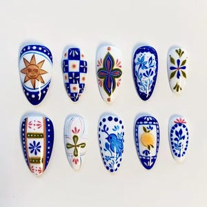 May include: A set of ten almond-shaped press-on nails featuring a variety of hand-painted designs in blue, white, yellow, and green. Patterns include a sun face, checkered squares, floral motifs, a small blue bird, and a lemon illustration.