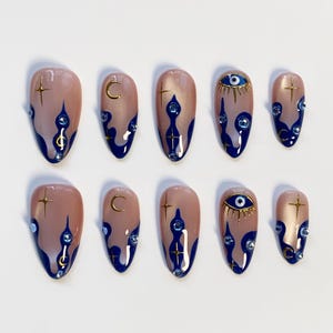 May include: Set of ten almond-shaped false nails. Each nail is painted a nude color with a dark blue flame design at the base. Some nails feature gold crescent moons, stars, or an eye design with gold accents and blue gems.