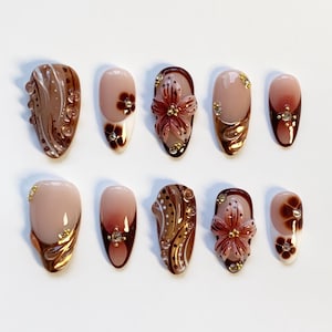 Op de afbeelding: Een set van tien kunstnagels in diverse ontwerpen. De nagels tonen een combinatie van nude, bruine en gouden kleuren. Sommige nagels hebben bloemversieringen en accenten, terwijl andere een gradiënteffect hebben.
