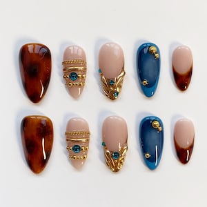 May include: A set of ten artificial nails in various designs. The nails feature a tortoiseshell pattern, nude bases with gold and teal accents, and navy blue with gold embellishments. The nails are oval-shaped.