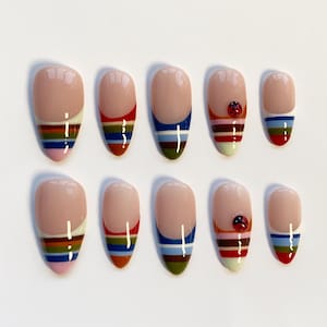 May include: A collection of ten almond-shaped false nails. Each nail has a nude base with a colourful striped design at the tip. Some nails feature a small ladybird accent. The stripes include red, blue, green, yellow, and pink.