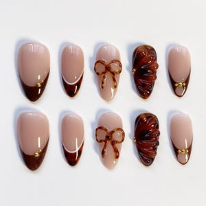 Peut inclure: Un ensemble de dix faux ongles en forme d'amande. Les ongles présentent divers motifs, notamment une base nude avec une pointe marron foncé, un trait blanc, un nœud marron et un motif texturé marron foncé. Certains ongles ont des détails dorés.