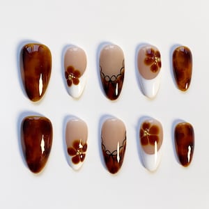 Könnte beinhalten: Ein Set von zehn ovalen Kunstnägeln. Die Designs umfassen einfarbiges Braun, Blumenmuster mit goldenen Akzenten und zweifarbige Nägel mit weißen Spitzen und braunen Blumendetails. Die Nägel sind auf einer weißen Oberfläche angeordnet.