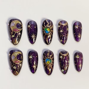 Könnte beinhalten: Ein Set aus zehn lila Press-on-Nägeln mit Himmelsmotiven. Jeder Nagel hat goldene Akzente, darunter Monde, Sterne und Sonnen, mit einem zentralen Juwel. Einige Nägel haben kleine, klare Strasssteine. Die Nägel sind oval.