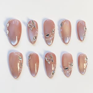 May include: A set of ten artificial nails in a light pink hue, adorned with silver-colored rhinestones and butterfly accents. The nails have an oval shape and a glossy finish, perfect for a special occasion or everyday wear.