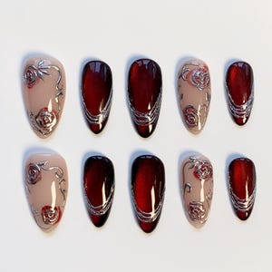 May include: A set of ten artificial nails. The nails feature a combination of designs, including a nude base with silver rose accents and deep red with silver swirls. The nails are oval-shaped and arranged on a white surface.
