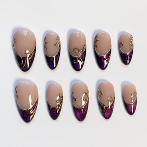 May include: A set of ten almond-shaped false nails. Each nail has a nude base, a deep purple glitter accent at the base, and gold filigree detailing. The nails are displayed on a white surface.