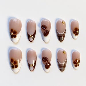 May include: A set of ten almond-shaped false nails. The nails feature a variety of designs, including a white tip with a brown flower, a brown and nude gradient, and a brown plaid pattern. Some nails have coffee bean accents.
