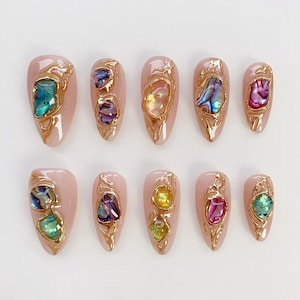 May include: A set of ten almond-shaped artificial nails. Each nail is painted a light pink and decorated with a gold-coloured metal frame. The frames hold colourful, iridescent stones in shades of blue, purple, yellow, and pink.