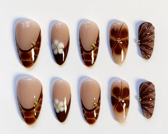 Puede incluir: Un conjunto de diez uñas artificiales con varios diseños. Algunas uñas presentan una base nude con estampado de cocodrilo marrón y detalles dorados. Otras tienen un diseño de flor blanca o un patrón de remolino marrón. Una uña tiene un diseño texturizado y acanalado.