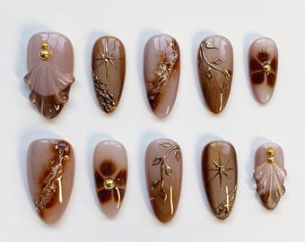 Puede incluir: Un conjunto de diez uñas artificiales en tonos marrones y rosas. Algunas uñas presentan detalles dorados, incluyendo diseños florales, estrellas y pequeñas cuentas doradas. Las uñas son de forma ovalada y están dispuestas sobre una superficie blanca.