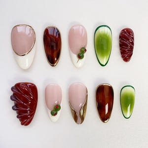 May include: A collection of ten almond-shaped false nails. The designs include solid colours like pink, brown, and green, and decorative elements such as gold trim and small green embellishments. Some nails have a textured, sculpted appearance.