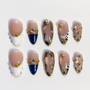 May include: A set of ten artificial nails in various designs. Some nails are nude with gold accents, others feature gold and blue details, and some have a black and white pattern. Several nails are adorned with small, light blue gemstones.