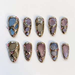 Puede incluir: Juego de diez uñas artificiales con un marco de metal dorado y un diseño abstracto colorido. Las uñas presentan tonos de azul, morado y rosa, con pequeñas gemas rosas. Las uñas tienen forma ovalada.