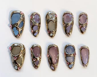 May include: Set of ten artificial nails with a gold-colored metal frame and a colorful, abstract design. The nails feature shades of blue, purple, and pink, with small pink gems. The nails are oval-shaped.