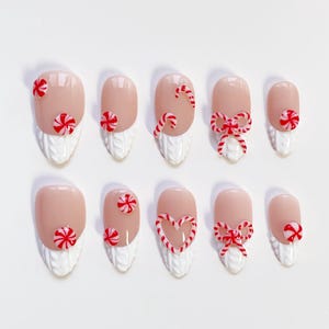 May include: A set of ten artificial nails with a light pink base, decorated with red and white candy cane and bow designs. The lower portion of each nail features a white, textured knit pattern, creating a festive holiday theme.