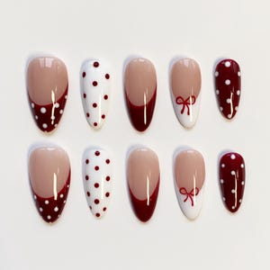 Puede incluir: Un conjunto de diez uñas postizas en forma de almendra. Las uñas presentan una variedad de diseños, incluyendo una base nude con una punta burdeos y lunares blancos, una base blanca con lunares burdeos y un lazo burdeos.