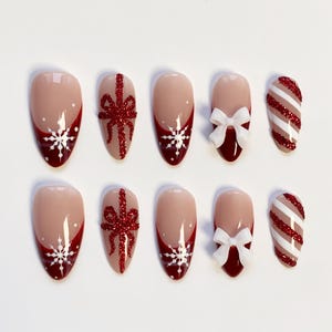 Puede incluir: Un conjunto de diez uñas artificiales con un diseño festivo. Las uñas presentan una base nude con detalles en rojo, copos de nieve blancos, lazos rojos con purpurina y patrones a rayas rojas y blancas. Las uñas son ovaladas.