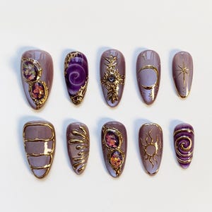 Pode incluir: Um conjunto de dez unhas posti&ccedil;as lavanda com enfeites dourados. Os desenhos incluem espirais douradas, pedras preciosas, um sol, uma lua e uma estrela. As unhas s&atilde;o ovais e dispostas sobre uma superf&iacute;cie branca.