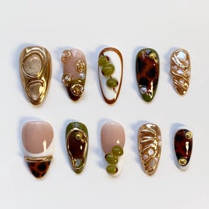 May include: A collection of ten artificial nails in various designs. The nails feature a range of colors, including gold, brown, green, and white, with embellishments like pearls and abstract patterns. The shapes vary, showcasing a modern nail art aesthetic.