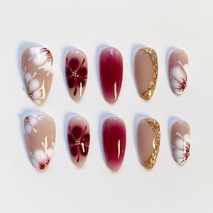 Pode incluir: Um conjunto de dez unhas artificiais com vários designs. Algumas unhas são nude com desenhos florais brancos e detalhes dourados. Outras apresentam um gradiente vermelho escuro ou nude com linhas decorativas douradas. As unhas são ovais.