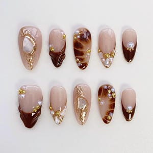May include: A collection of ten almond-shaped false nails. The nails have a nude base with brown tips and are adorned with gold and pearl details. Some nails have a marbled effect, while others feature ornate gold designs.
