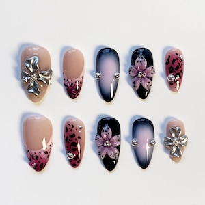 May include: A set of ten artificial nails in various designs. Some nails feature a nude base with a dark pink leopard print, while others have a gradient effect from nude to black. Embellishments include silver bows, flower accents, and small silver studs.