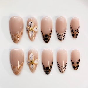 Pode incluir: Um conjunto de dez unhas postiças em forma de amêndoa. As unhas apresentam uma base nude com vários designs, incluindo uma estampa de leopardo, listras de tigre pretas e uma flor 3D com detalhes dourados e bolinhas brancas.