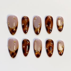 May include: Set of ten almond-shaped false nails. Each nail features a gradient design with a nude base, transitioning to a dark brown tip with gold glitter accents. Small gold studs are evenly spaced across each nail.