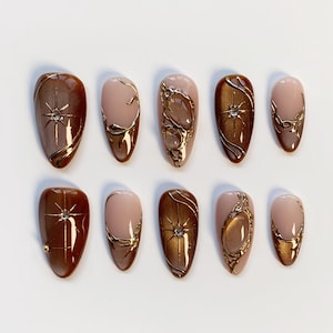 May include: A collection of ten almond-shaped false nails. The nails are in shades of brown, nude, and gold. Some have starburst and swirl patterns, while others feature gold detailing. The nails are displayed on a white background.