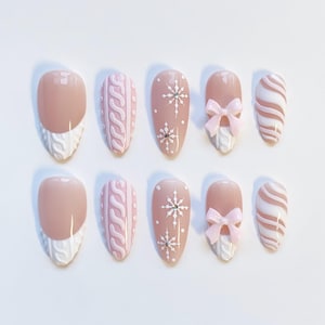 Könnte beinhalten: Ein Set aus zehn mandelförmigen Kunstnägeln. Die Nägel weisen eine Vielzahl von Designs auf, darunter eine hellrosa Basis mit weißen Zopfmuster-Akzenten, Schneeflockenmuster und rosa Schleifen. Einige Nägel haben eine nude Basis mit weißen Zopfmuster-Spitzen.
