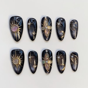 Puede incluir: Un conjunto de diez uñas postizas negras y doradas. Cada uña presenta diseños celestiales, incluyendo soles, estrellas y lunas crecientes, con acentos iridiscentes y de pedrería. Las uñas son de forma ovalada y tienen un acabado brillante.