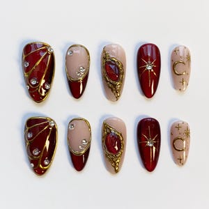 May include: A collection of ten artificial nails in diverse designs. The nails showcase a deep red, nude, and gold colour scheme. Some nails have gold embellishments, rhinestones, and celestial designs, while others are solid colours.