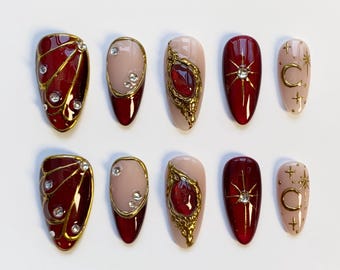 Puede incluir: Un conjunto de diez uñas artificiales con varios diseños. Las uñas presentan una combinación de colores rojo oscuro, nude y dorado. Algunas uñas tienen adornos dorados, pedrería y diseños celestiales, mientras que otras son de un solo color.