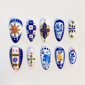 May include: A set of ten almond-shaped press-on nails featuring hand-painted designs in blue, white, yellow, and green. Patterns include a sun face, blue checkered squares, floral motifs, a small blue bird, and a yellow lemon within a blue border.