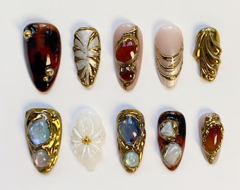 Puede incluir: Una colección de diez uñas artificiales ornamentadas. Cada uña presenta un diseño único, que incluye filigrana dorada, piedras preciosas y detalles de perlas. Los colores varían desde carey hasta nude, con tonos dorados y joya.