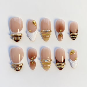 May include: A set of ten artificial nails in various designs. The nails feature a nude base with brown ombre and gold accents. Some nails have gold rings with rhinestones, while others have gold studs and white decorative elements.