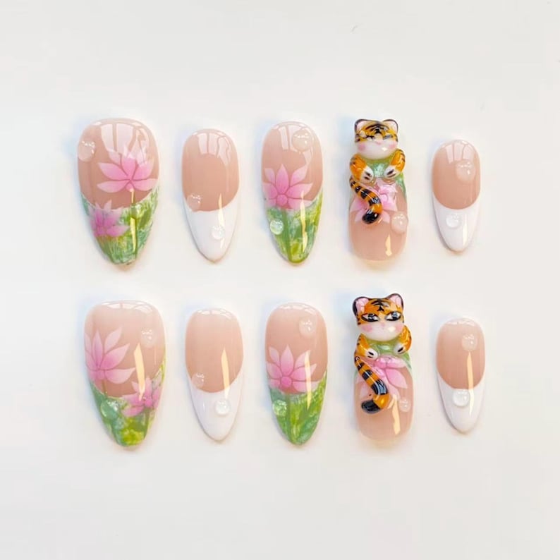 May include: A set of ten almond-shaped false nails. The nails feature a variety of designs, including pink lotus flowers, white tips, and a 3D tiger figurine. The nails are painted with a glossy finish.