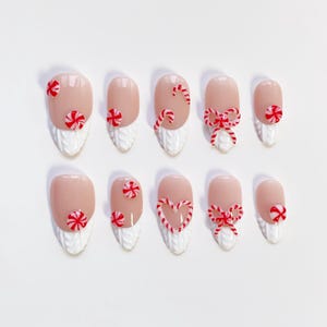 May include: A set of ten artificial nails with a light pink base and white textured tips. Each nail is decorated with red and white candy cane designs, including candy cane swirls, bows, and peppermint candies. The nails are arranged on a white surface.