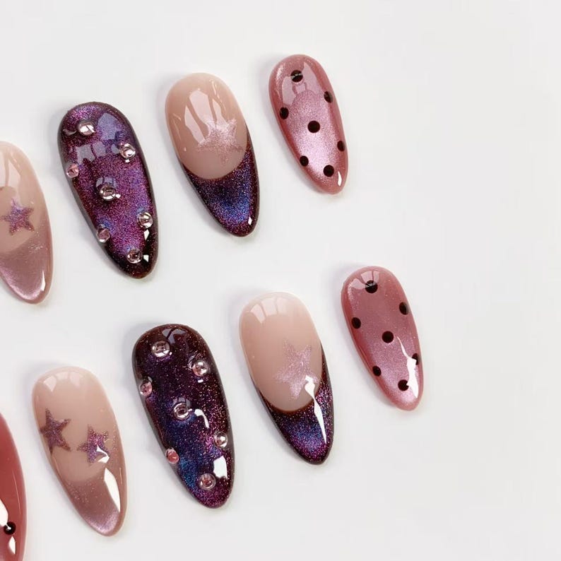 May include: A collection of artificial nails in various designs. Some nails are a light pink with a star design, others have a dark purple glitter effect with rhinestones, and some are a light pink with a dark purple tip. One nail has black polka dots.