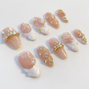 May include: A collection of artificial nails in various designs. The nails feature a nude pink base with white, gold, and rhinestone accents. Some nails have a lace pattern, while others have floral or pearl details. The nails are oval-shaped.