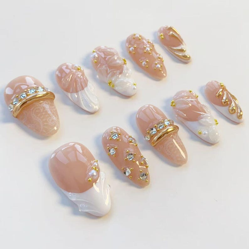 May include: A collection of artificial nails in various designs. The nails feature a nude pink base with white, gold, and rhinestone accents. Some nails have a lace pattern, while others have floral or pearl details. The nails are oval-shaped.