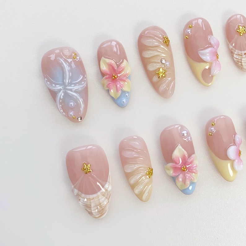 May include: A collection of artificial nails with various designs. Some nails feature a light pink base with floral accents in pink, yellow, and blue. Others have seashell patterns or gold embellishments. The nails are oval-shaped.